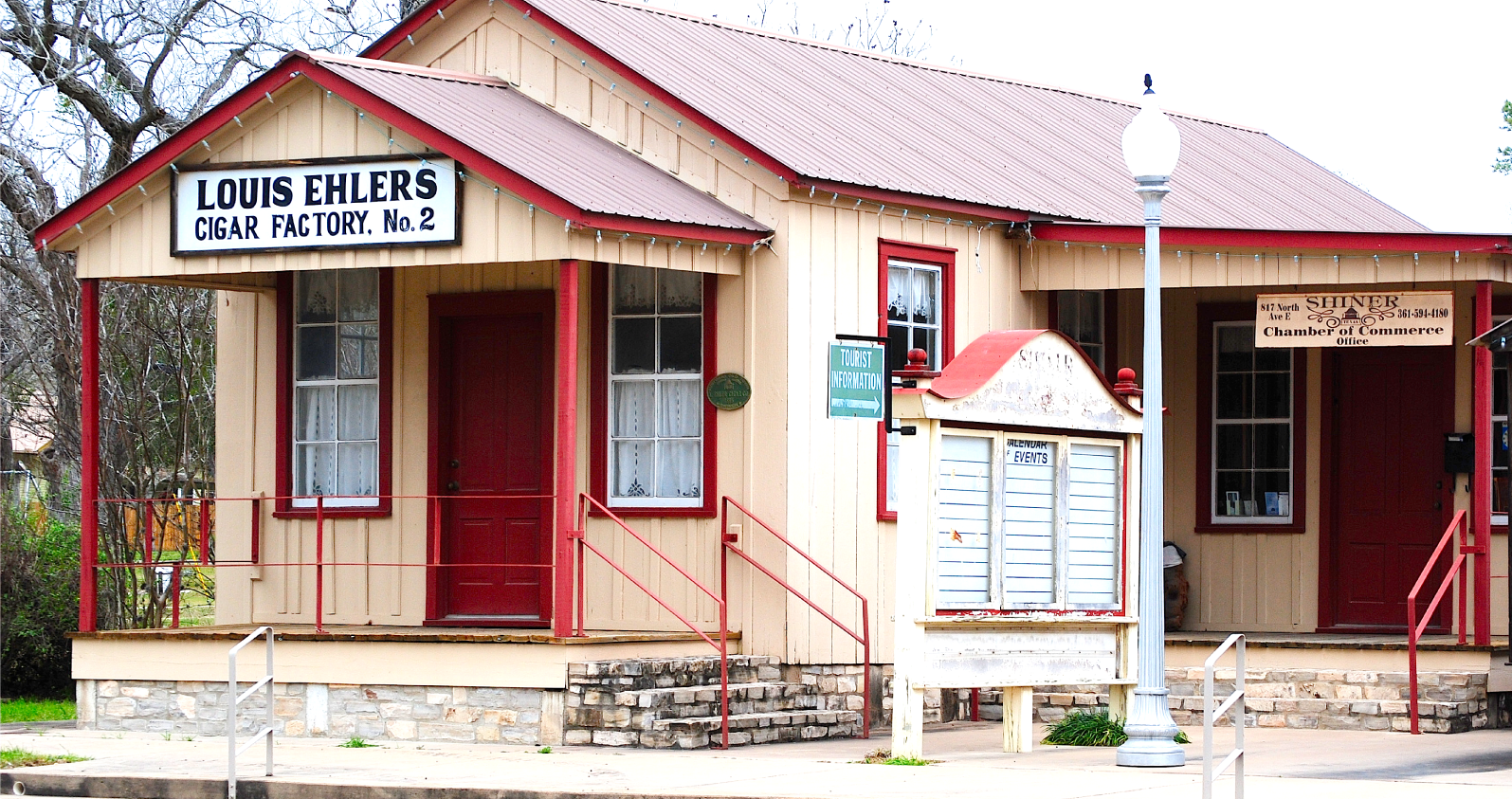 Photo of the historic Louis Ehlers Cigar Factory No. 2, now serving as the Shiner Chamber of Commerce with a tourist information center. The building is painted beige with red trims, featuring a front porch and a stone foundation.
